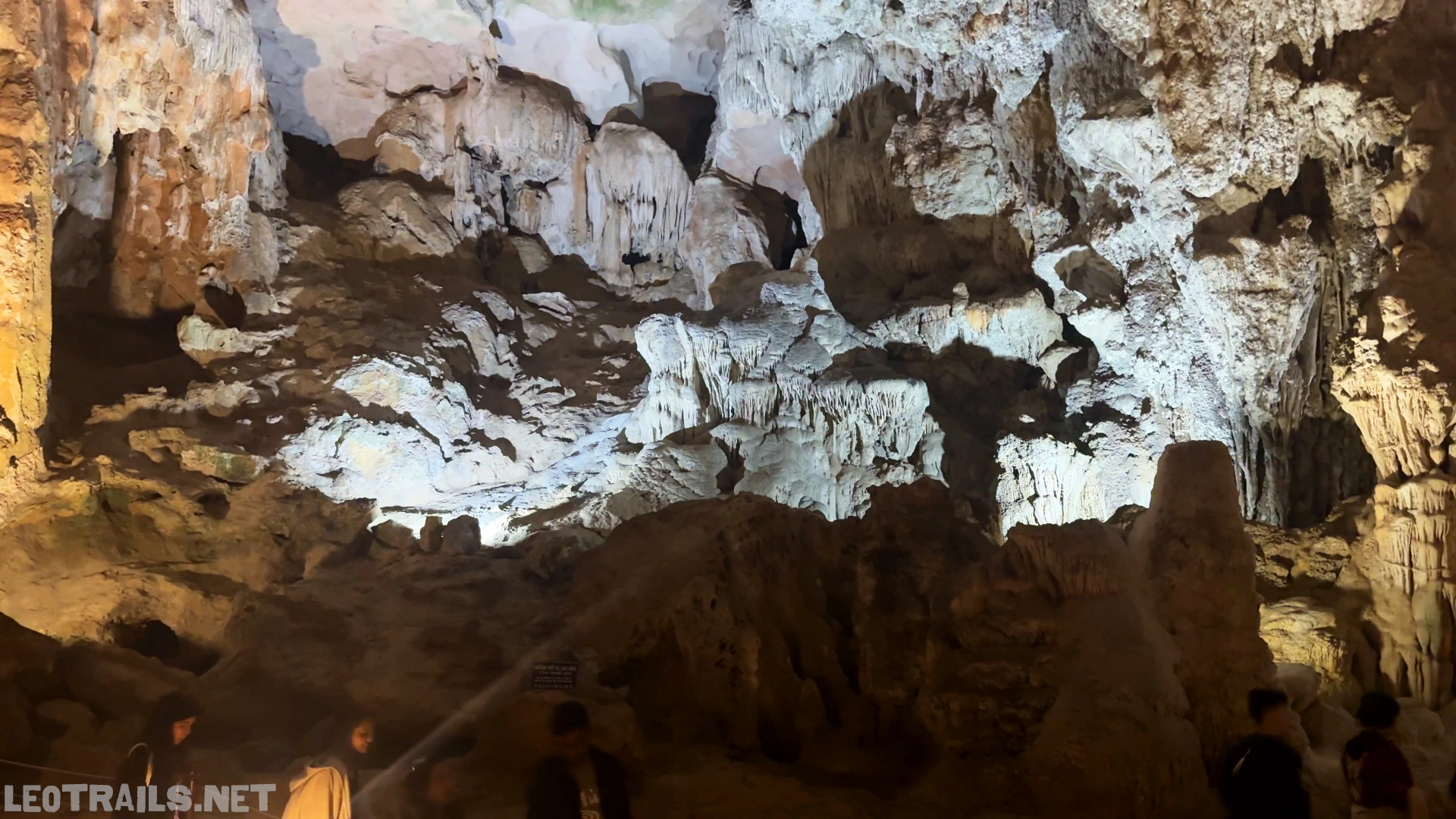 Interior close-up view of a massive rock chamber inside Sung Sot Cave in Ha Long Bay, Vietnam, showcasing heavily textured stalactite columns and multi-layered limestone formations illuminated by dramatic white and warm yellow spotlighting, with the silhouettes of tourists visible at the bottom.