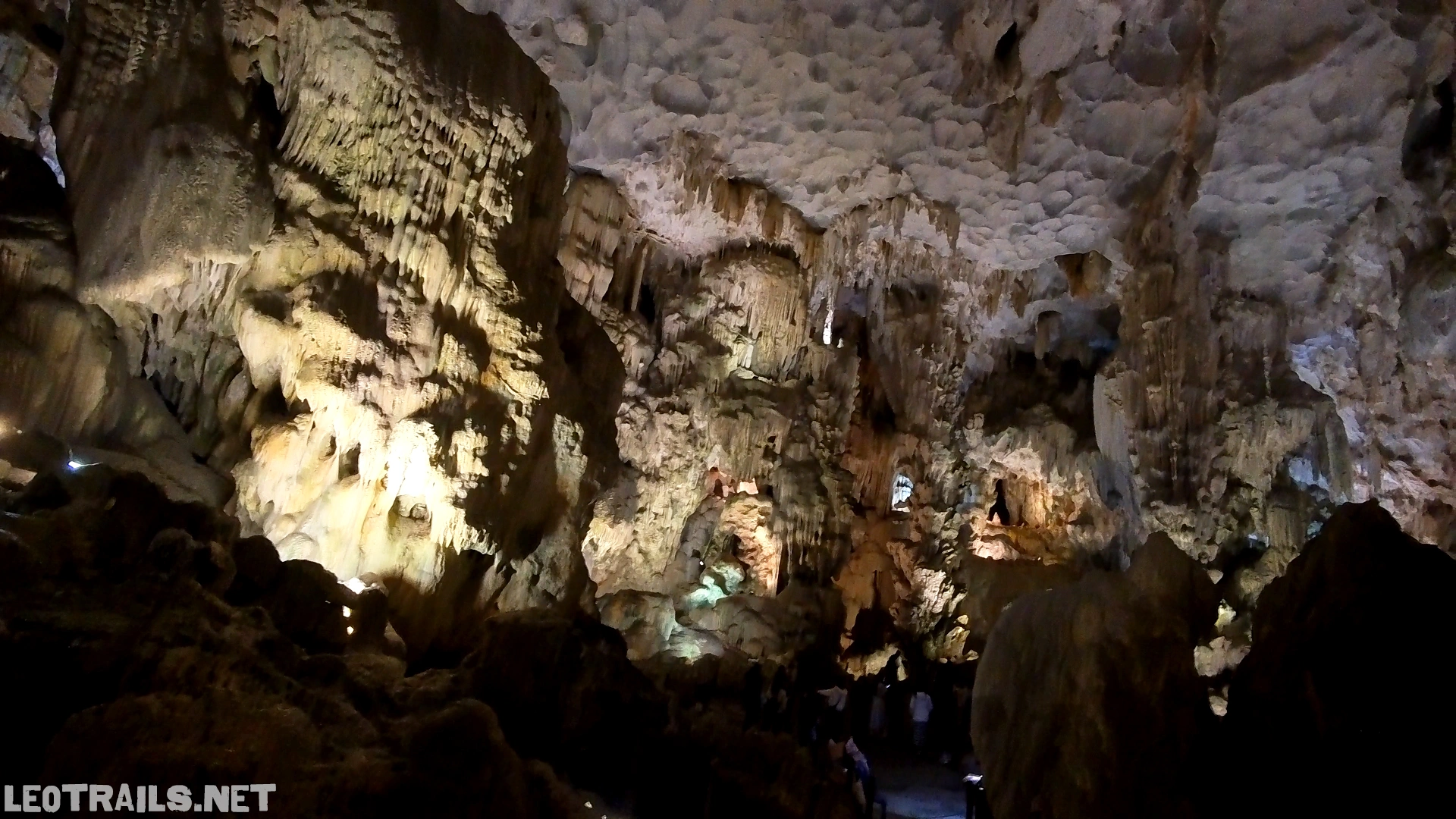 Wide-angle interior view of a massive underground chamber inside Thien Cung Cave in Ha Long Bay, Vietnam. The image displays a vast textured ceiling with undulating, cloud-like limestone patterns illuminated by soft ambient lighting, overlooking a deeply shadows-cast lower cavern floor filled with complex stalagmite rock formations.