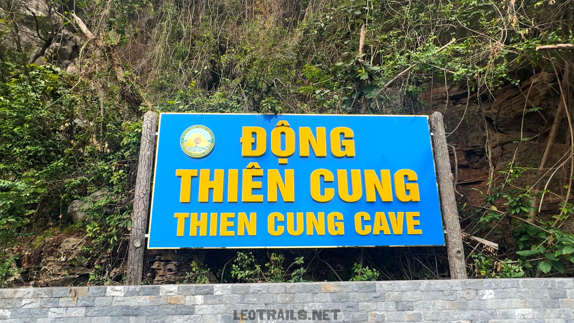 A bright blue sign with yellow text reading "ĐỘNG THIÊN CUNG - THIEN CUNG CAVE" surrounded by green foliage at the entrance area.