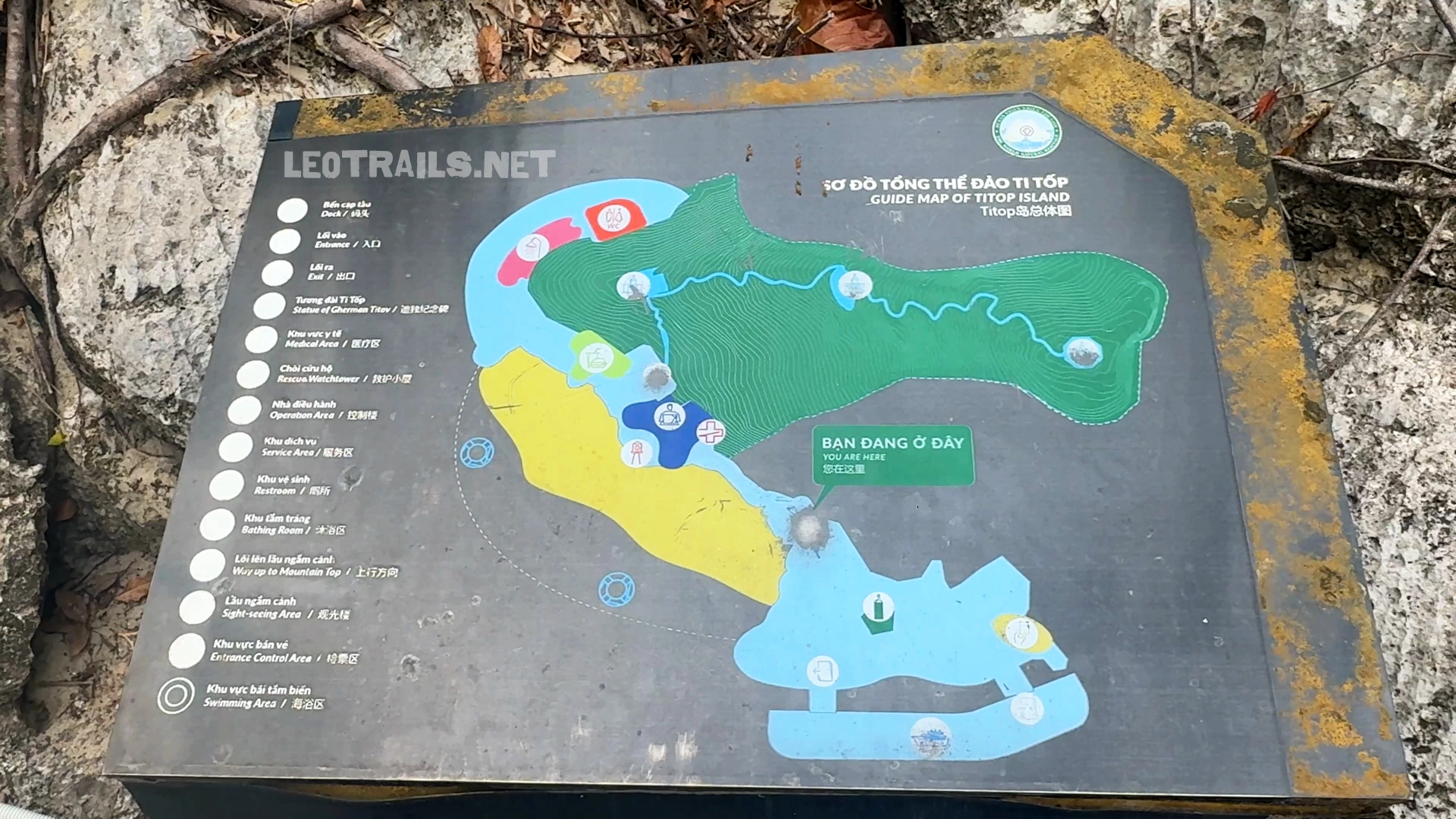 Close-up shot of a weathered outdoor tourist signboard showing the colorful "Guide Map of Titop Island" in Ha Long Bay, Vietnam. The black metal sign displays a green and yellow illustrated map of the island's geography, hiking trails, viewing deck, and crescent swimming beach, set against a natural limestone rock background.