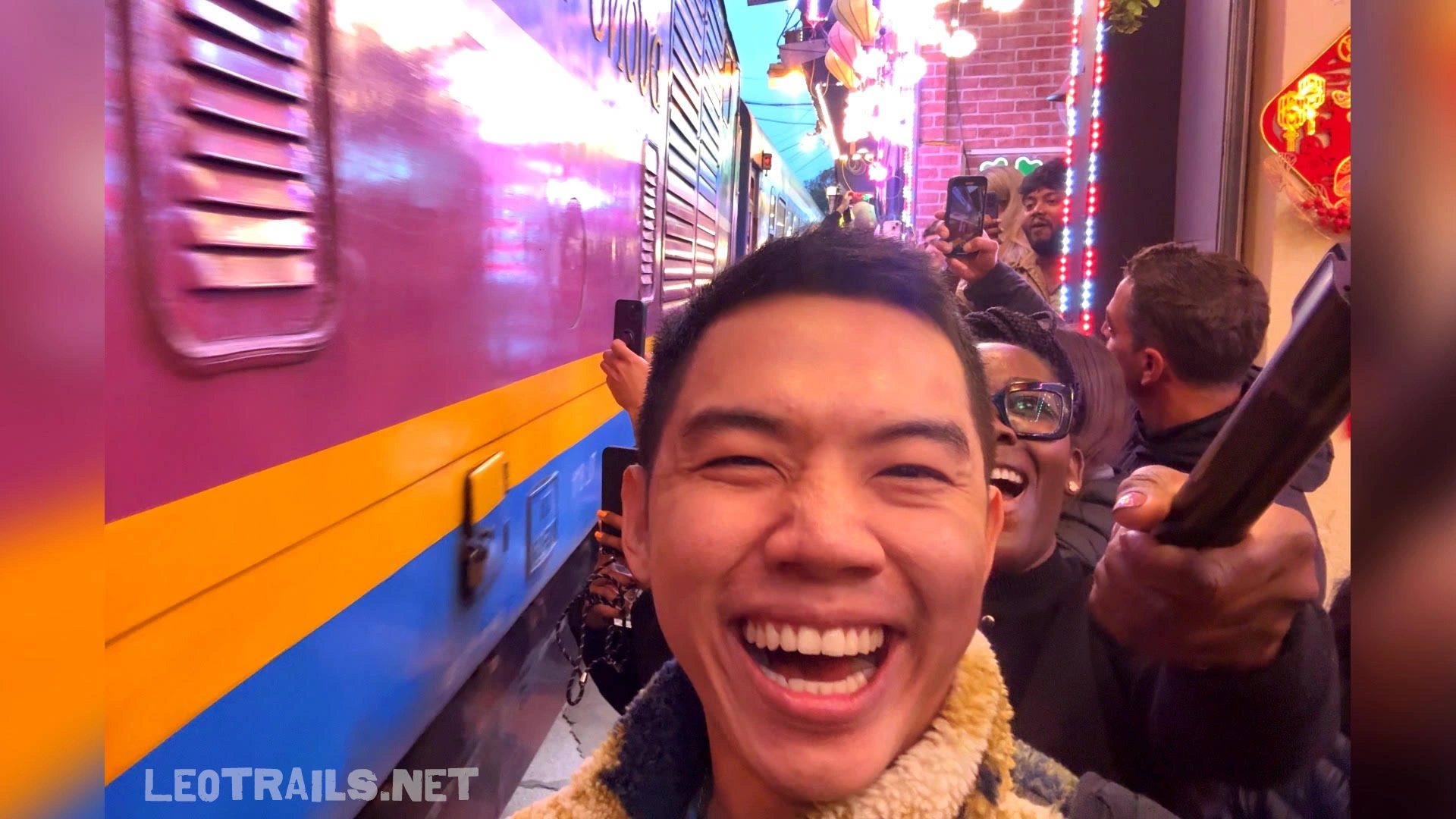 A laughing man takes a close-up selfie as a purple, yellow, and blue train passes immediately behind him next to a crowd of excited onlookers at night.