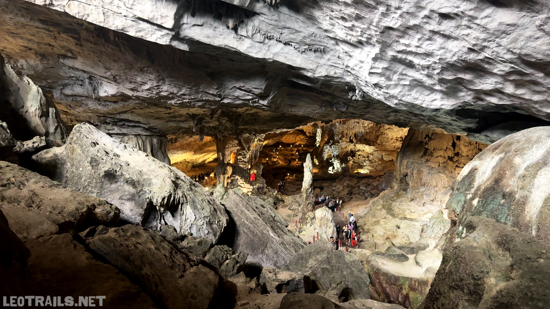 Interior view of the massive Sung Sot Cave (Surprise Cave) in Ha Long Bay, Vietnam, showing a vast underground limestone cavern with soaring rock ceilings, dramatic stalactite formations, and small groups of tourists walking along a paved path illuminated by golden ambient lighting.