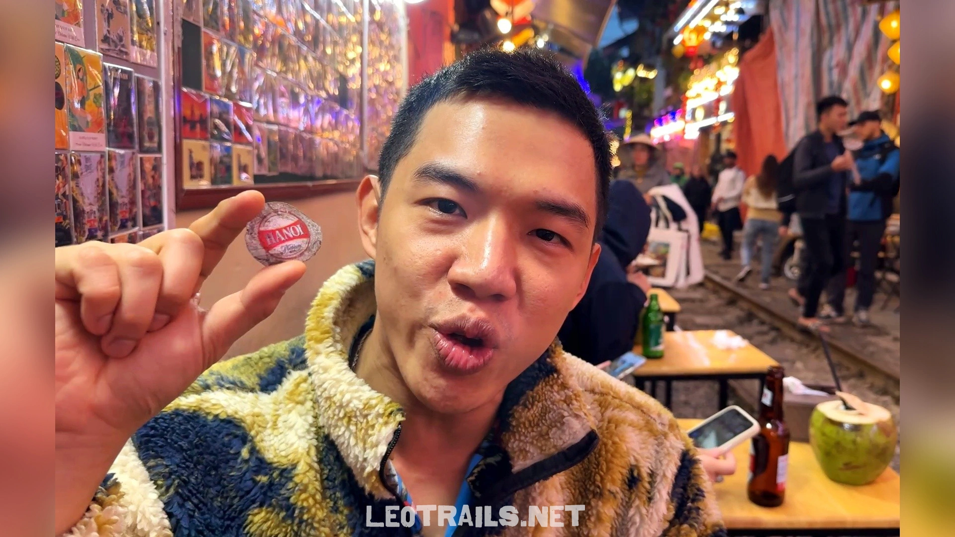 A close-up selfie of a man holding a silver and red Hanoi Beer bottle cap while sitting at a trackside table at Hanoi Train Street.