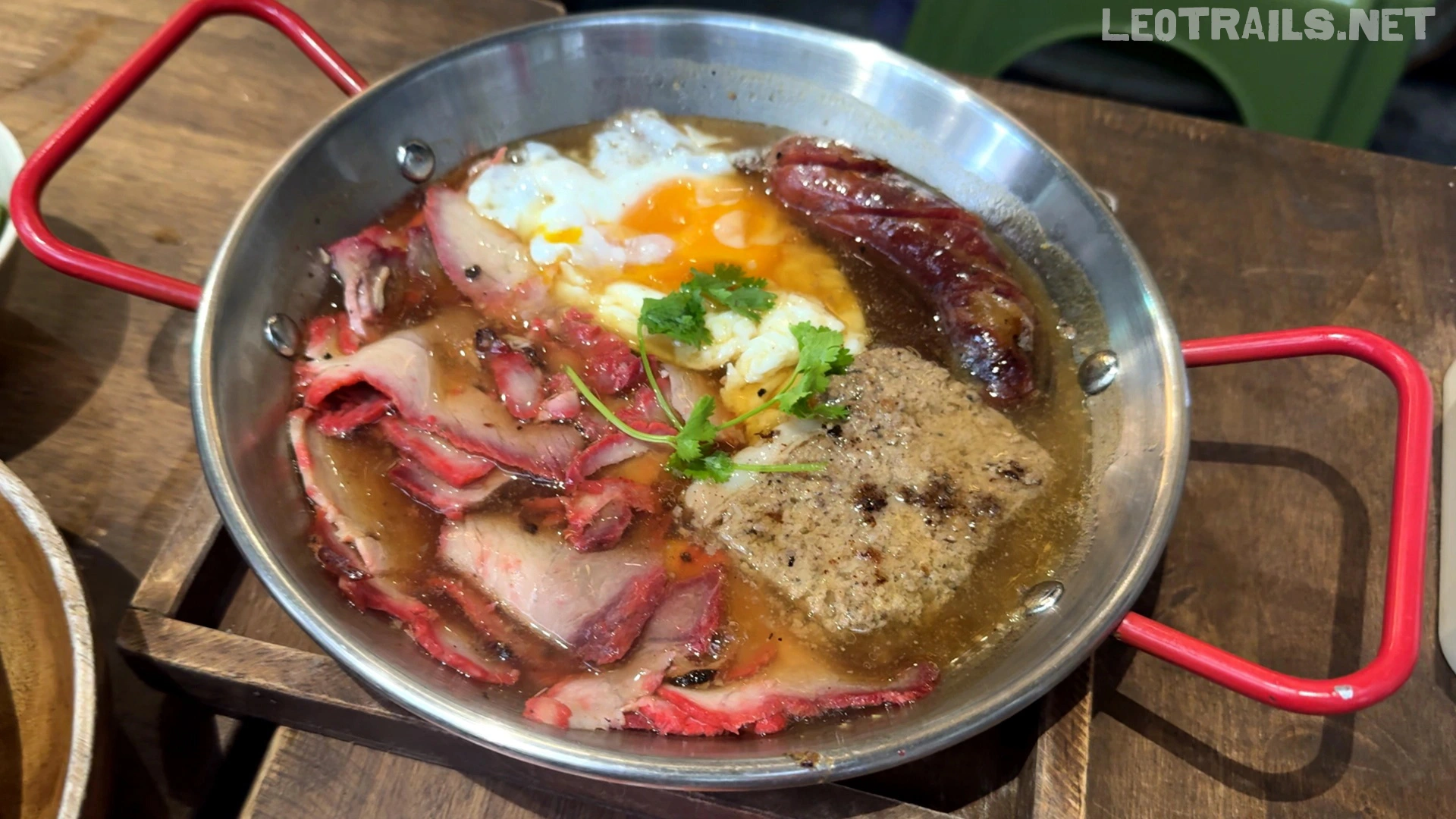 A stainless steel skillet with red handles containing Vietnamese Bánh Mì Chảo, featuring sliced char siu pork, a fried egg with runny yolk, a sausage, and a square slab of pâté in a rich savory sauce.