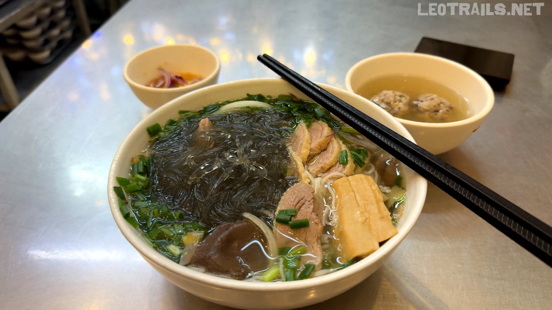 A white bowl of Vietnamese miến ngan (combination of rice vermicelli and glass noodles with duck meat) topped with sliced duck meat, bamboo shoots, mushrooms, and green onions, served with a side bowl of broth containing meatballs.