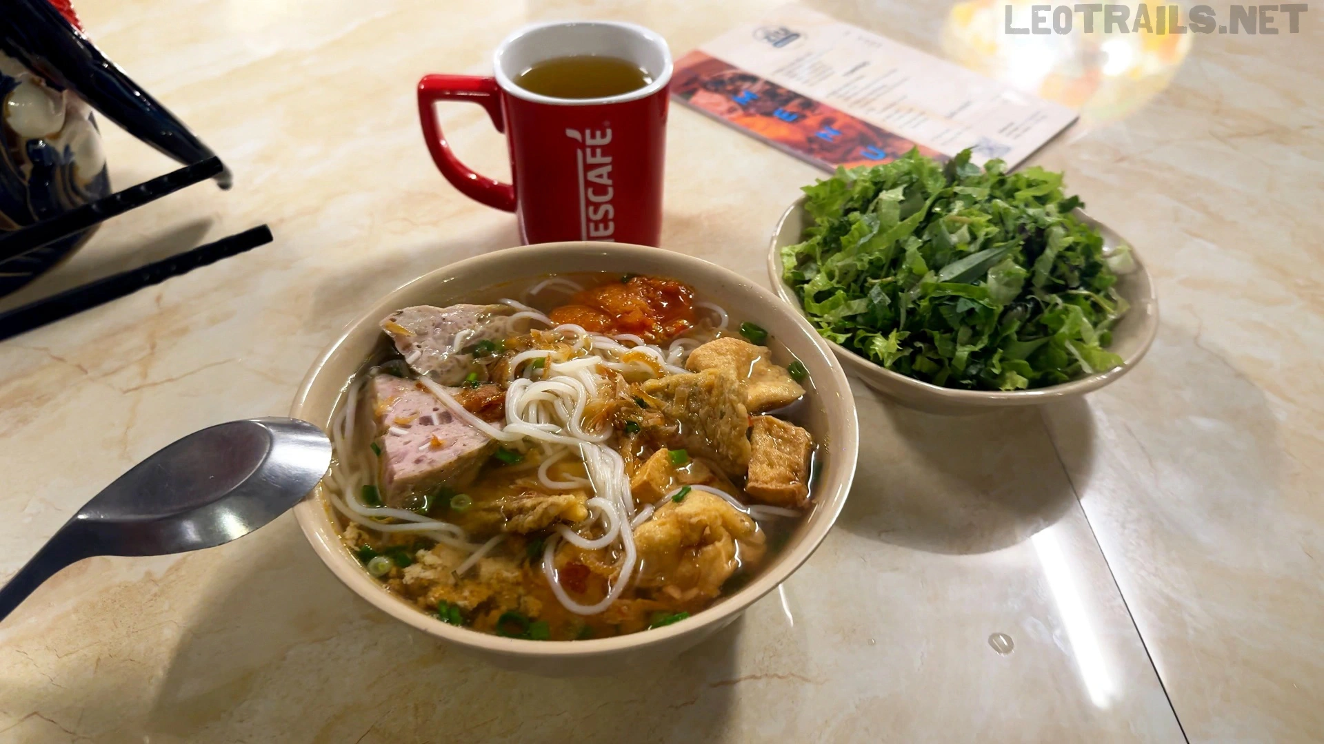 A tan bowl of Vietnamese bún riêu (crab paste noodle soup) containing rice vermicelli, fried tofu blocks, and pork sausages, served on a table next to a bowl of fresh shredded greens and a red Nescafé mug.