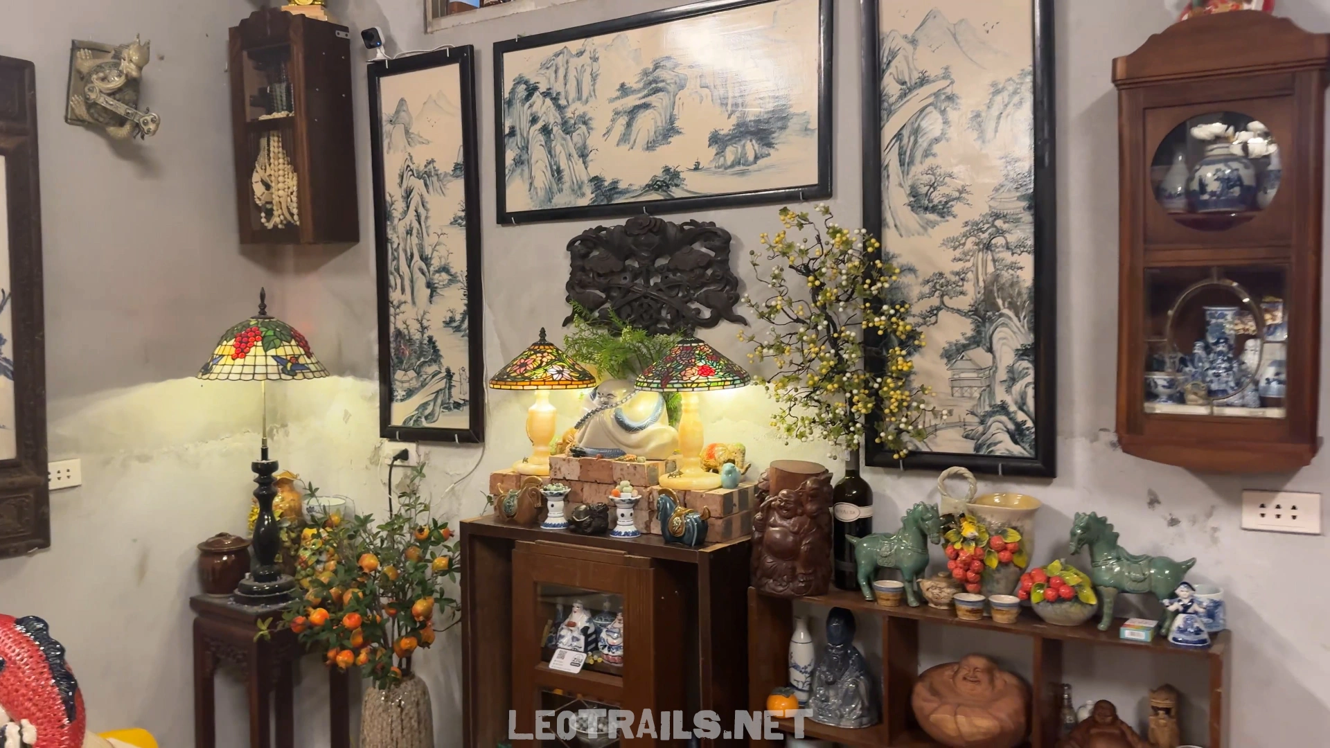 A wall display inside Bún Ziu Gánh restaurant featuring traditional Vietnamese and Chinese ceramic art pieces, wooden display cabinets, tiffany-style table lamps, and blue and white porcelain landscape paintings.
