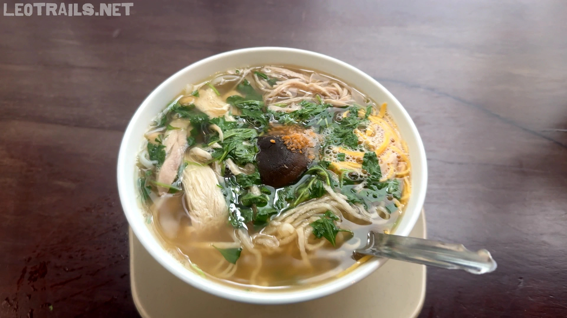 A white bowl of traditional Vietnamese bún thang rice vermicelli soup containing shredded chicken, sliced egg omelet, mushrooms, and fresh herbs in a clear broth on a dark wooden table.