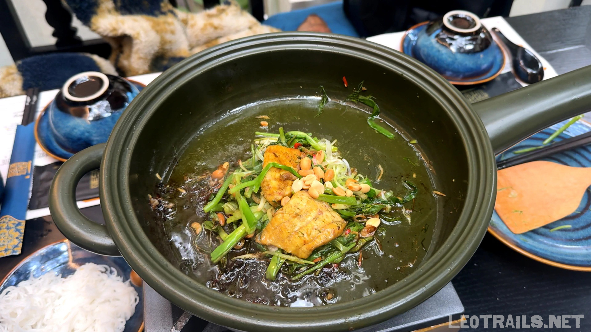 A green pan containing Vietnamese chả cá (turmeric fish with dill and green onions) topped with roasted peanuts, served with small bowls and rice vermicelli.