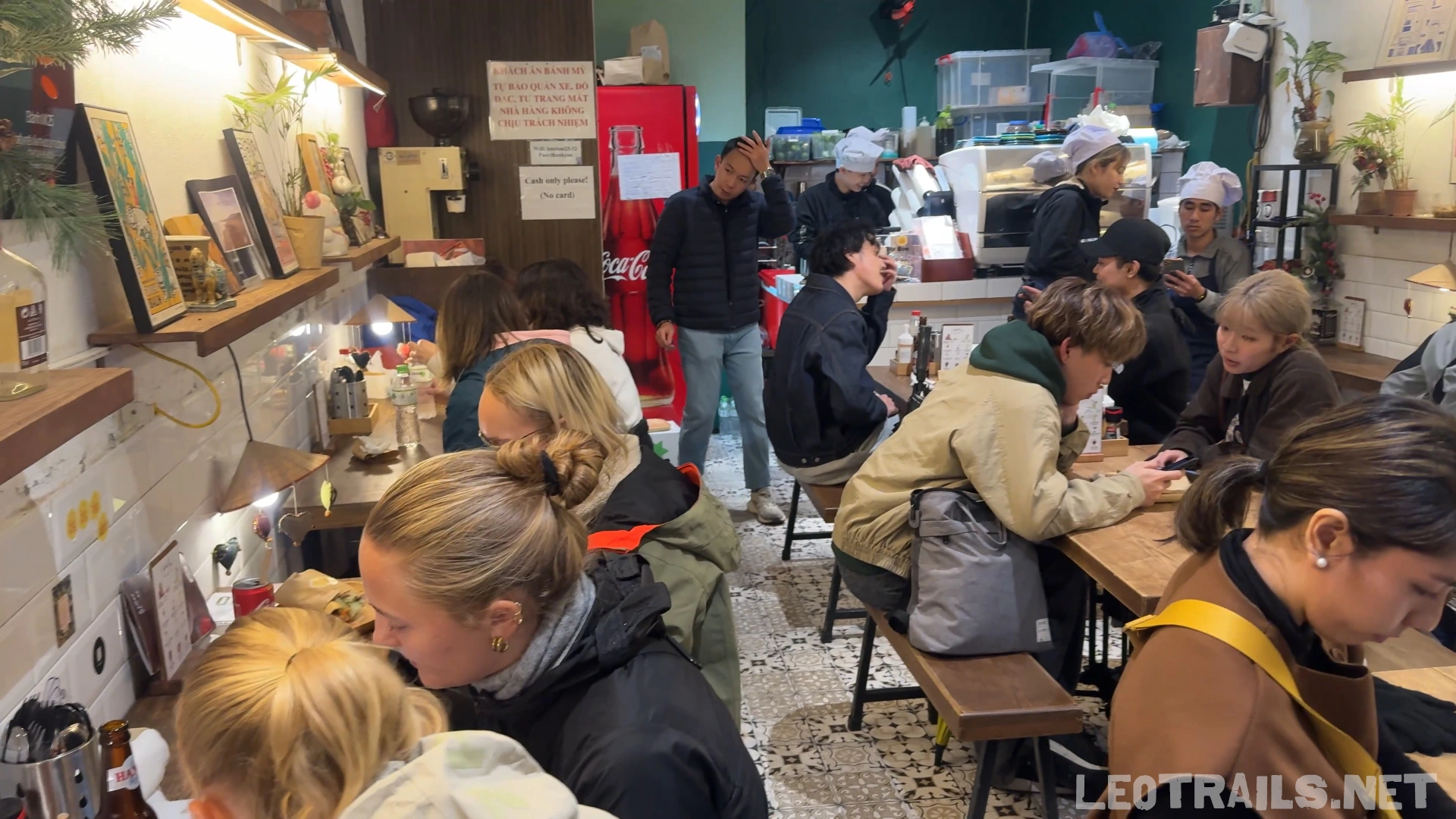  The crowded indoor dining room of Banh Mi 25 restaurant, showing local and international customers sitting at wooden tables and benches with kitchen staff working in the background.