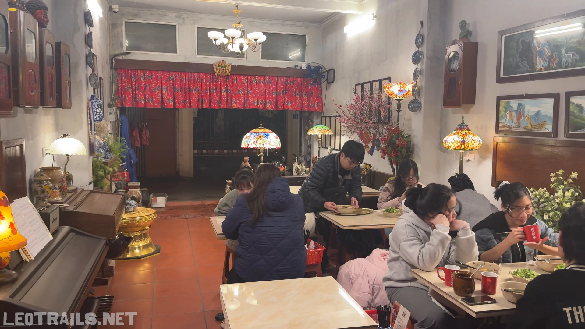 The indoor dining room of Bún Ziu Gánh restaurant in Hanoi, featuring customers sitting at tables, tiffany-style glass lamps, red floral curtains, framed artwork, and an old piano on the left.