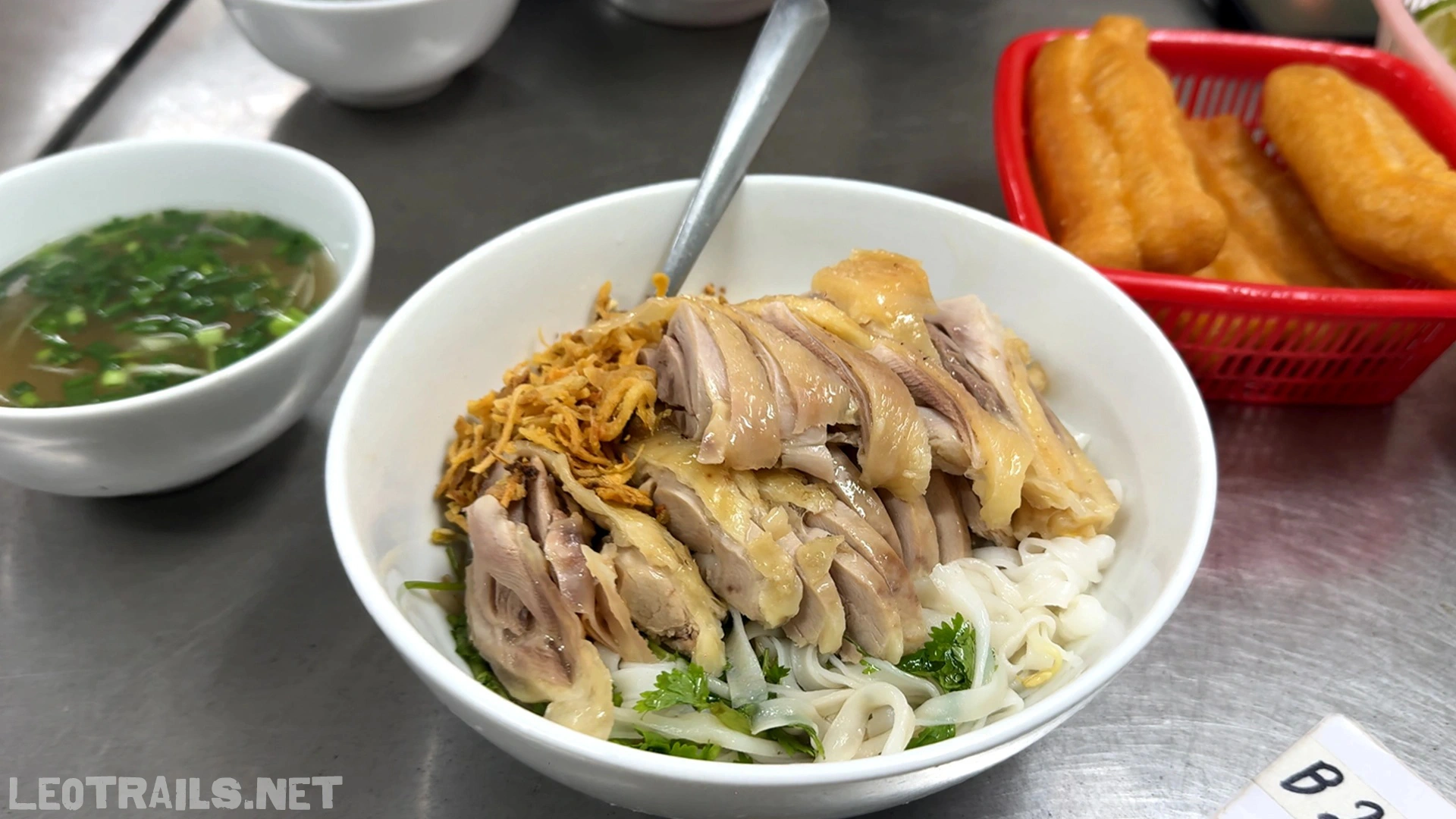 A white bowl of Vietnamese phở gà trộn (dry chicken pho) topped with sliced steamed chicken skin-on, fried shallots, and cilantro, served with a side bowl of broth and a red basket of quẩy (fried dough sticks).
