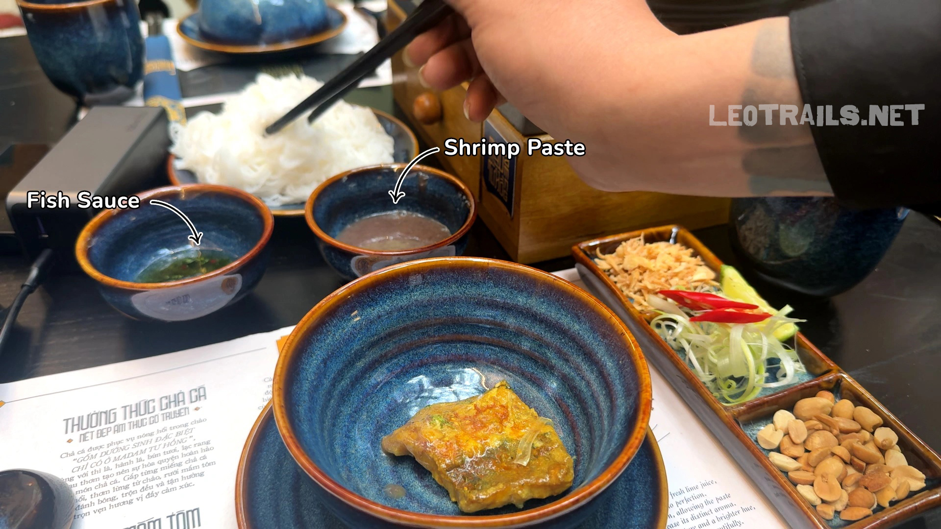 A table setting for Vietnamese Chả Cá showing a bowl with a piece of grilled turmeric catfish, surrounded by small side bowls labeled fish sauce and shrimp paste, fresh herbs, peanuts, and rice vermicelli.