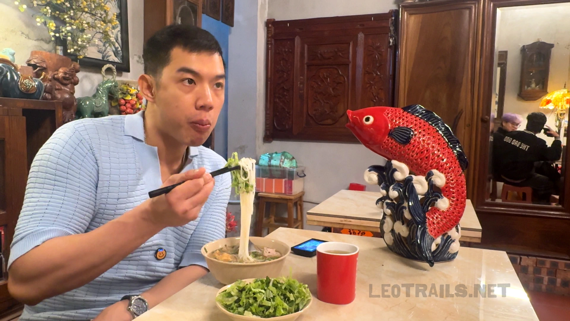 A man in a light blue textured polo shirt using chopsticks to lift rice vermicelli noodles from a bowl of bún riêu at a restaurant table next to a large red ceramic fish statue.