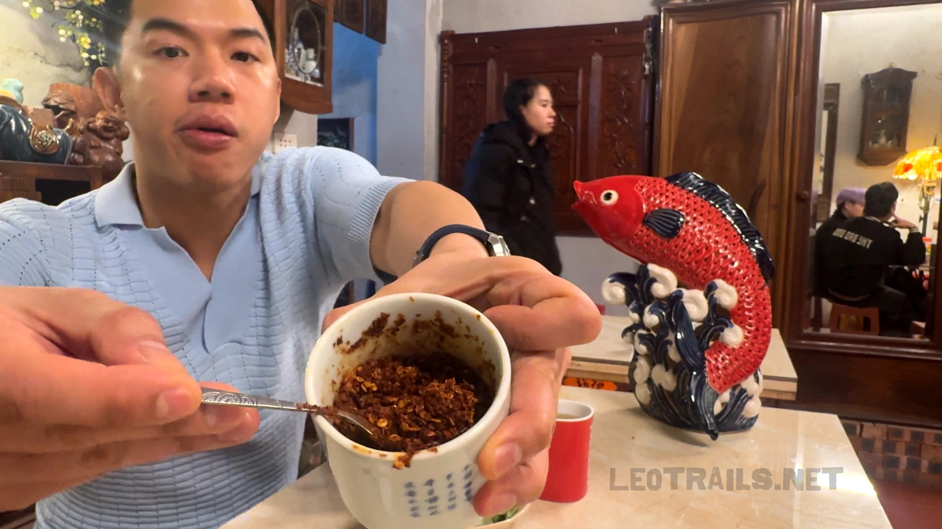 A man holding up a small white ceramic cup filled with dark red roasted chili flakes (ớt chưng) with a small metal spoon, showing the table condiment close up at Bún Ziu Gánh.