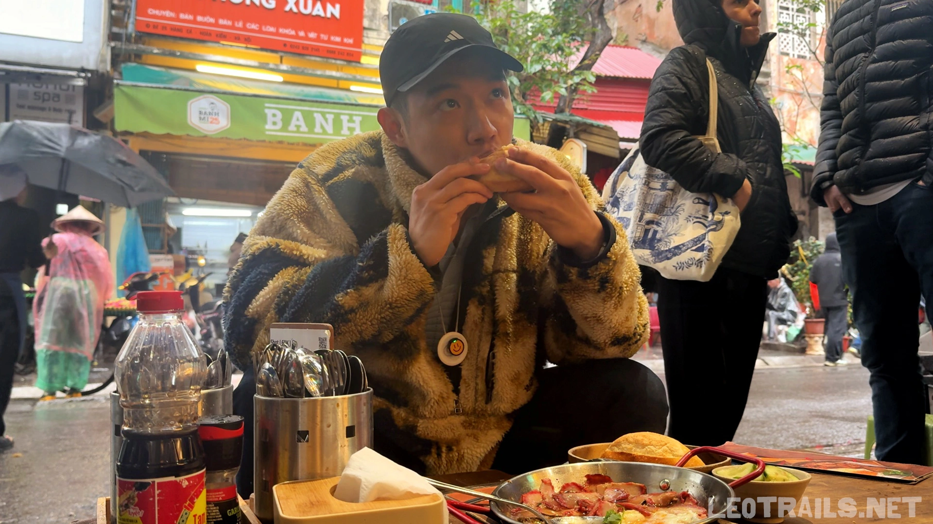 A man wearing a patterned fleece jacket and black cap eating a crispy banh mi sandwich at a street food table, with a sizzling Bánh Mì Chảo skillet platter and the Banh Mi 25 storefront visible behind him.