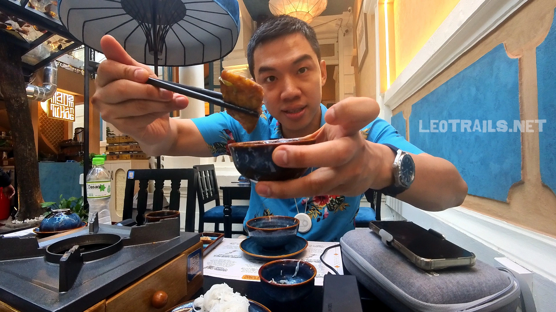 A man wearing a blue floral shirt using chopsticks to dip a piece of turmeric fish (Chả Cá) into a small sauce bowl at a well-lit restaurant table.