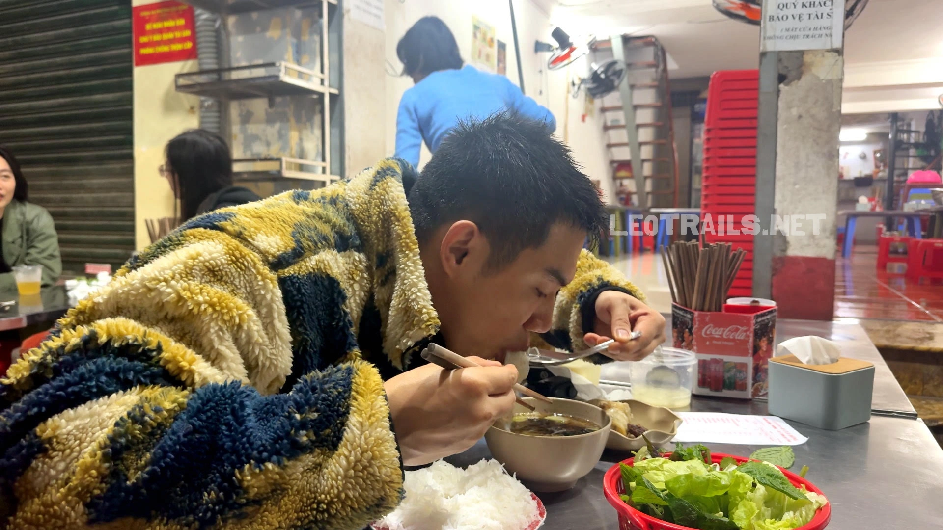 A man wearing a fuzzy patterned jacket eating bún chả with chopsticks at a local street restaurant table filled with fresh herbs, noodles, and dipping broth.