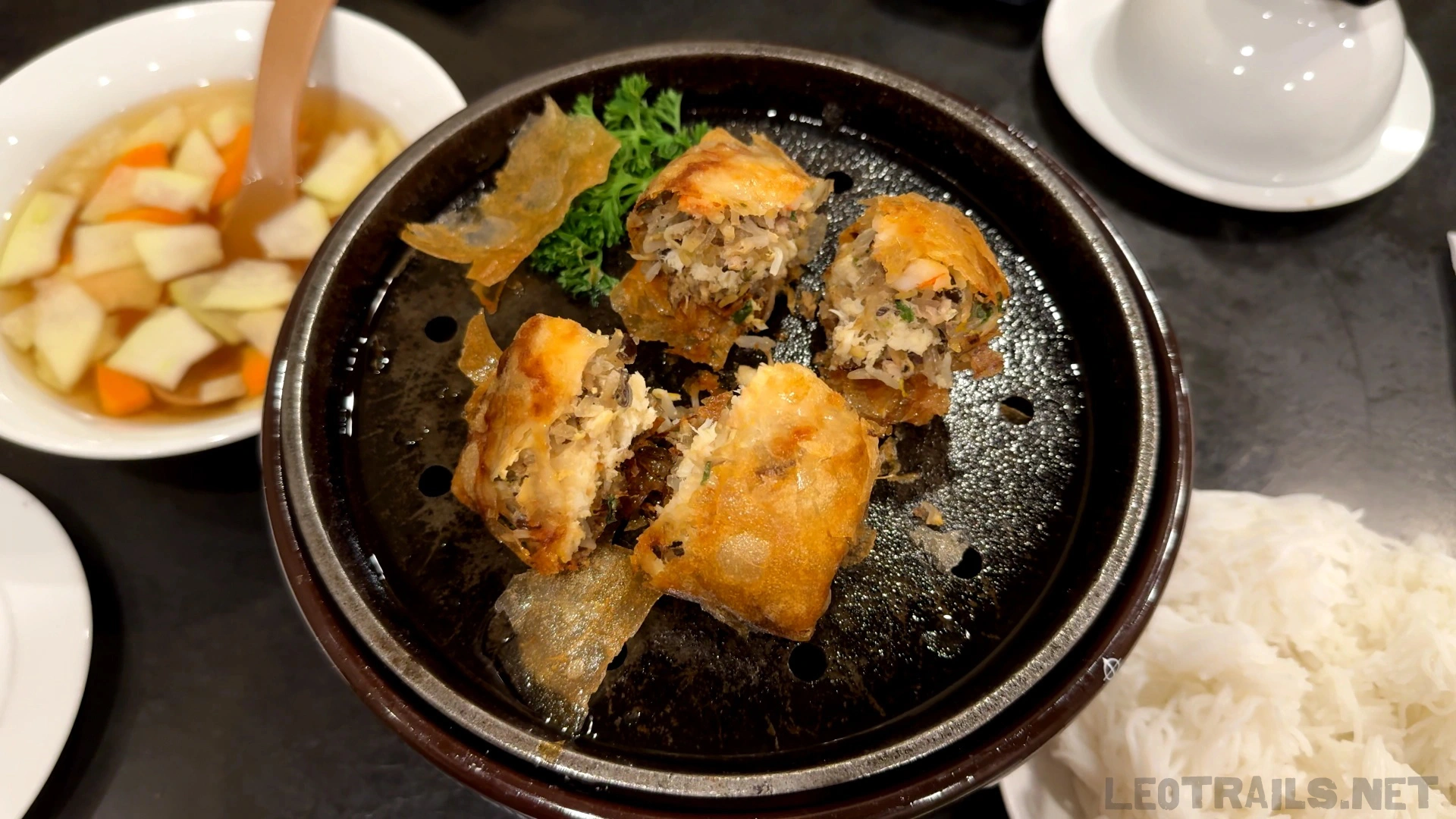 Crispy Vietnamese fried spring rolls (Nem Rán) cut into pieces and served on a black grill pan, accompanied by a bowl of sweet and sour dipping sauce with pickled vegetables and a side of rice vermicelli.
