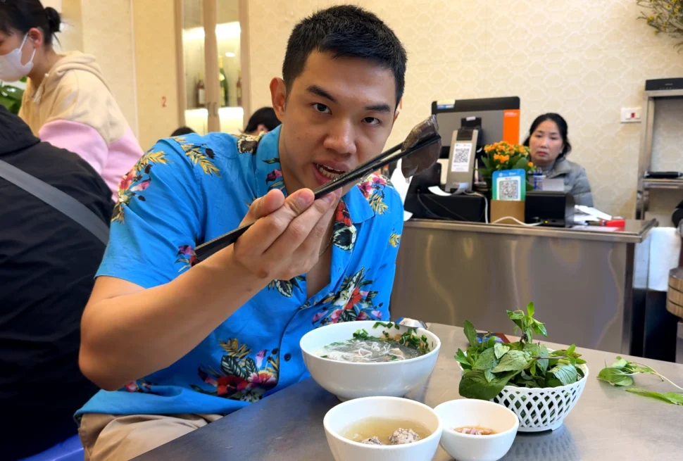 A man wearing a bright blue floral shirt holding a piece of meat with black chopsticks above a white bowl of noodle soup, next to a bowl of fresh green herbs.