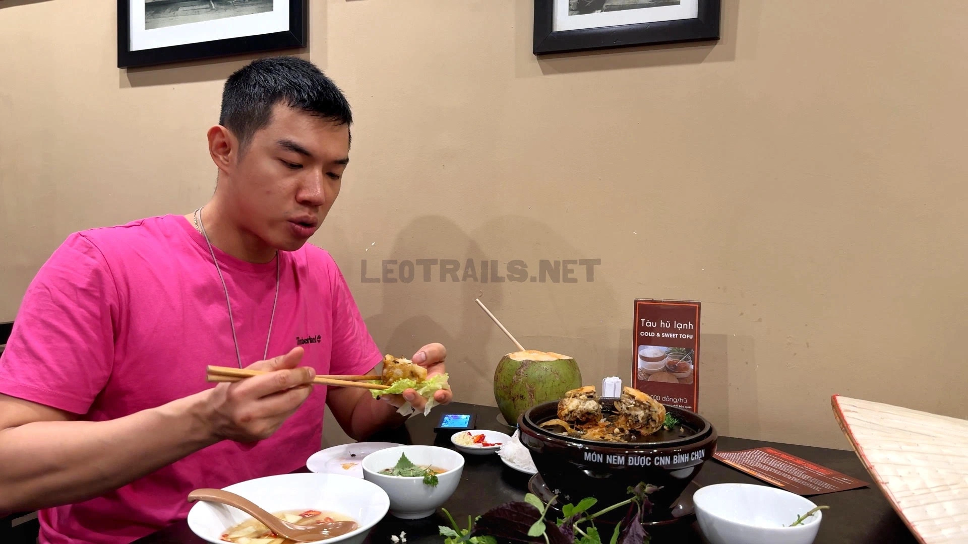 A man wearing a bright pink t-shirt using chopsticks to wrap a crispy piece of crab spring roll with fresh lettuce at a restaurant table featuring a coconut drink and small dipping bowls.